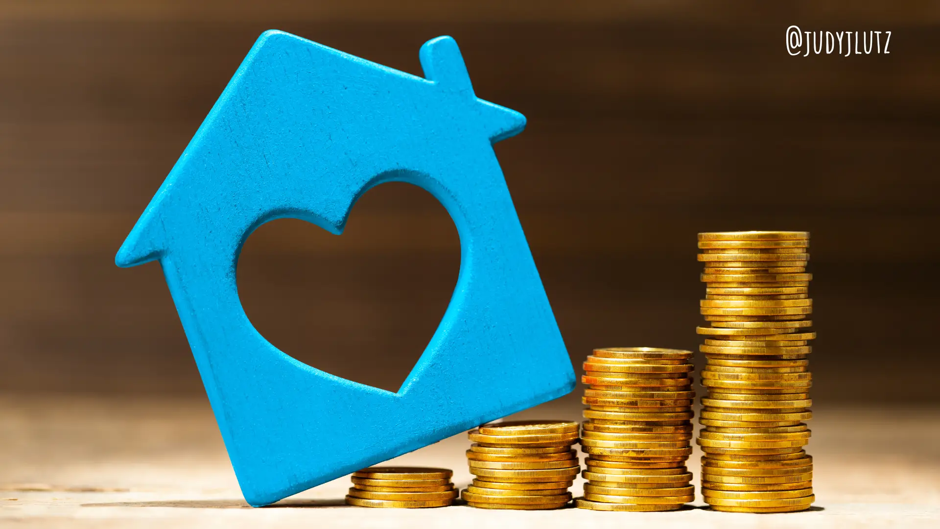 a blue house with a heart shape and stacks of gold coins on a wooden background indicating monitored home security systems can be expensive.